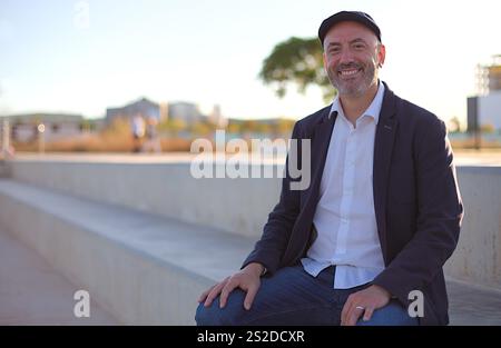 Ein fröhlicher Mann mittleren Alters in lässiger Kleidung sitzt auf einer Betonbank in einer hellen urbanen Umgebung im Freien, mit einer verschwommenen Stadtlandschaft im Hintergrund Stockfoto