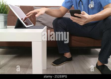 Medizin und Gesundheit Zusammenhang Arzt Hand arbeiten mit smart-Phone, digitale Tablet-Computer, Stethoskop, sitzen auf dem Sofa im Wohnzimmer Stockfoto