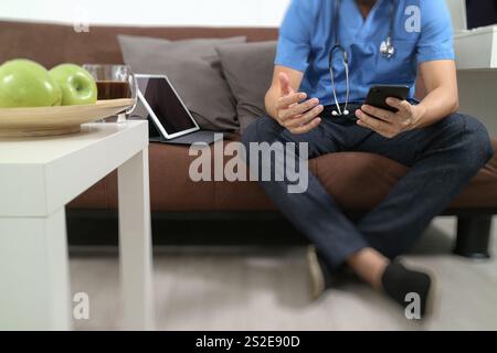 Medizin und Gesundheit Zusammenhang Arzt Hand arbeiten mit smart-Phone, digitale Tablet-Computer, Stethoskop, sitzen auf dem Sofa im Wohnzimmer Stockfoto