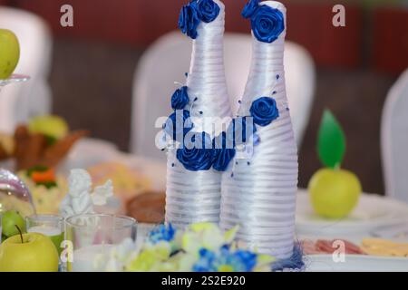 Zwei festliche Champagnerflaschen sind mit künstlichen blauen Rosenblüten verziert. Auf dem festlichen Hochzeitstisch. Stockfoto