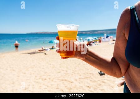 Eine Frau hält am Strand einen Plastikbecher Bier. Es gibt noch mehrere andere Personen im Hintergrund. Selektives Schärfen Stockfoto