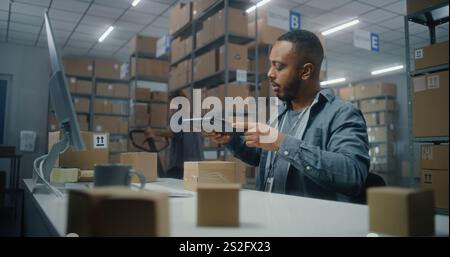 Afroamerikanischer Lagermitarbeiter scannt das Paket mit einem Tablet-Computer, schaut auf den PC-Bildschirm, arbeitet in der Post. Mitarbeiter des Sortierzentrums, der Kartons für den Versand transportiert. Speicherung von E-Commerce-Geschäften. Stockfoto