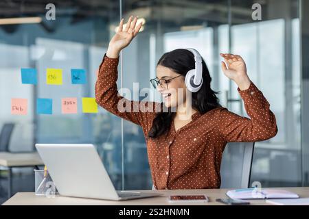Fröhliche Frau, die Musik am Arbeitsplatz im Büro hört. Geschäftsfrau mit Kopfhörern, die sich am Laptop entspannen, tanzen und singen, während sie am Tisch sitzt. Stockfoto