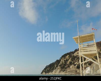 Aussichtsturm an leerem Strand aus hellem Holz mit roter und weißer Flagge, die an sonnigem Tag starken Wind vor dem Hintergrund der Klippe am Meer zeigt Stockfoto