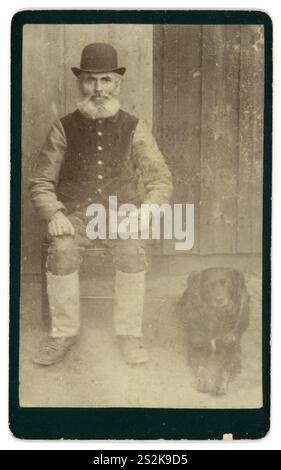 Original viktorianischer Carte de Visite (Visitenkarte oder CDV) viktorianischer Mann viktorianischer Gentleman. Der ältere Mann ist bärtig und aus dieser Kleidung ist ein Mann der Arbeiterklasse mittleren Alters, der einen Bowler-Hut und Lederleggings trägt, vor einem Bauernhaus sitzt, ein Buch hält, einen Arbeitshund an seiner Seite. Vielleicht ist der Mann ein Bauer oder Landarbeiter. War dieser Mann alphabetisch oder Analphabet - das Buch ist wahrscheinlich eine fotografische Requisite. Das Foto stammt von den 1870er bis Anfang der 1880er Jahre Stockfoto