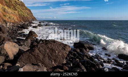 Ein malerischer Blick auf die Küste mit zerklüfteten Felsen und stürzenden Wellen gegen ein pulsierendes blaues Meer, mit einer grasbewachsenen Klippe unter klarem Himmel. Stockfoto