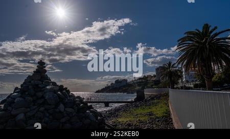 Eine Pyramide aus gestapelten Steinen an einem felsigen Strand unter einer hellen Sonne, mit Palmen, einem Pier und Gebäuden entlang der Küste im Hintergrund. Stockfoto
