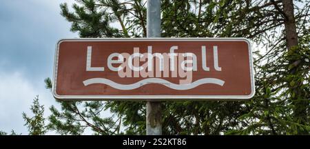 Braunes Lechfall-Wasserfall-Straßenschild in Füssen, Bayern, zwischen immergrünen Bäumen unter bewölktem Himmel. Stockfoto