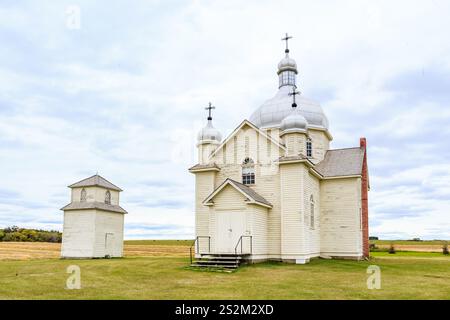 Eine kleine weiße Kirche mit einem hohen Turm sitzt auf einem Feld. Die Kirche ist von einigen kleinen Gebäuden umgeben. Der Himmel ist bewölkt und das Gras ist grün Stockfoto