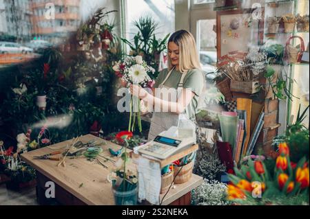 Floristin kreiert einen wunderschönen Blumenstrauß aus gemischten Blumen, während sie in einem Blumenladen arbeitet. Frau, die einen schönen Blumenstrauß auf der Co kreiert Stockfoto