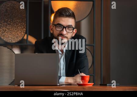 Porträt eines jungen glücklichen Geschäftsmannes, der an seinem Schreibtisch im Büro sitzt und in die Kamera schaut. Stockfoto