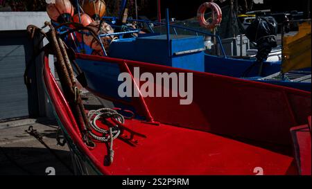 Ein leuchtendes rotes Fischerboot mit Seilen und Bojen, das neben blauen Booten in einem Hafen angedockt ist. Die kontrastierenden Farben schaffen eine markante nautische Szene. Stockfoto