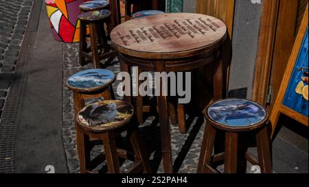 Ein hölzerner Tisch im Freien mit eingraviertem Text, umgeben von Hockern mit bunten bemalten Designs, auf einer kopfsteingepflasterten Straße. Stockfoto