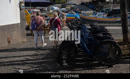 Eine Reihe von geparkten Fahrrädern auf einer kopfsteingepflasterten Straße in der Nähe farbenfroher Fischerboote, mit Fußgängern, die unter hellem Sonnenlicht in einer Küstenstadt spazieren. Stockfoto
