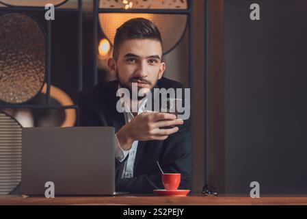 Porträt eines jungen glücklichen Geschäftsmannes, der an seinem Schreibtisch im modernen Büro sitzt und ein Telefon benutzt. Stockfoto