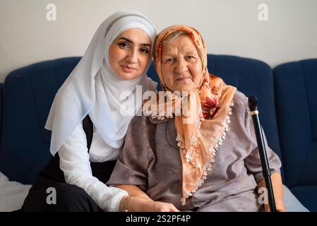 Porträt einer muslimischen alten Großmutter und einer erwachsenen Enkelin, die sich mit Liebe auf dem Sofa umarmt, während sie in die Kamera schauen. Stockfoto