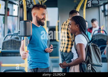 Ein junges multirassisches Paar, das in einem fahrenden Bus steht, während er redet und flirtet. Porträt von zwei verschiedenen lächelnden Freunden, die sich im Bus unterhalten, während sie zu W Stockfoto