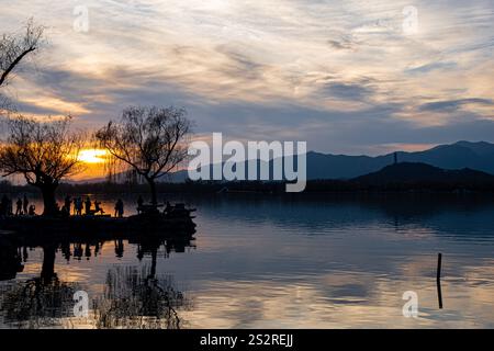 Während die goldene Sonne langsam über einem ruhigen See untergeht, steht eine wunderschöne Silhouette von Bäumen im Vordergrund, während majestätische Berge im Hintergrund aufsteigen Stockfoto