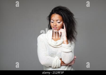 Durchdachtes weibliches Modemodell mit Kopf in der Hand, das vor grauem Hintergrund steht. Junge Dame mit geschlossenen Augen und Vitiligo Hautkrankheit ist im Studio. Stockfoto