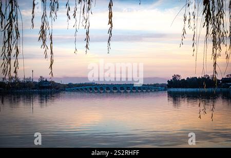 Eine atemberaubend schöne Brücke überspannt sich anmutig über einen ruhigen See bei einem malerischen Sonnenuntergang, mit einem großen Baum, der im Vordergrund steht Stockfoto