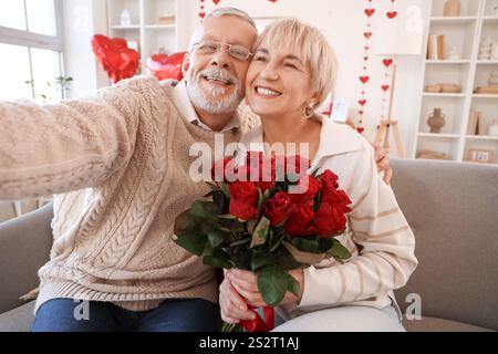 Glückliches reifes Paar mit einem Rosenstrauß, das zu Hause Selfie auf dem Sofa macht. Valentinstag-Feier Stockfoto