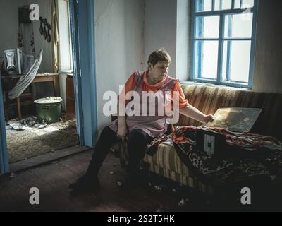 Irina in ihrem kleinen Haus an der Front, das größtenteils durch Beschuss zerstört wurde, heizt im Winter ein Zimmer mit kleinem Ofen, Kurahowka, Ukraine, Europ Stockfoto