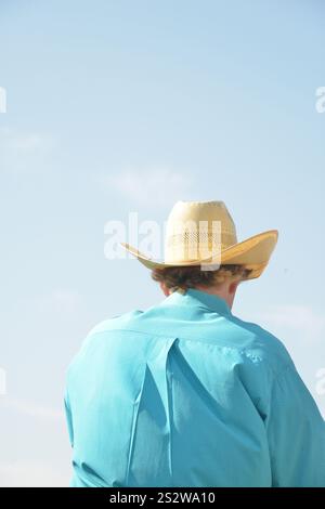 Ein Cowboy mit Strohhut und türkisfarbenem Hemd reitet an einem sonnigen Tag zu Pferd. Von hinten fotografiert, hält der Cowboy die Zügel. Stockfoto