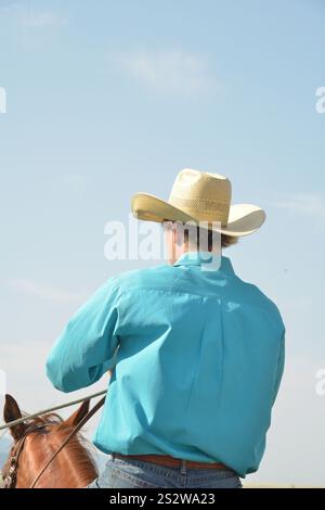 Ein Cowboy mit Strohhut und türkisfarbenem Hemd reitet an einem sonnigen Tag zu Pferd. Von hinten fotografiert, hält der Cowboy die Zügel. Stockfoto