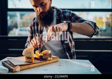 Attraktiver Hipster in kariertem Hemd, der in einem Restaurant einen veganen Burger isst. Kalorienreiche Speisen. Cheat-Mahlzeit. Stockfoto