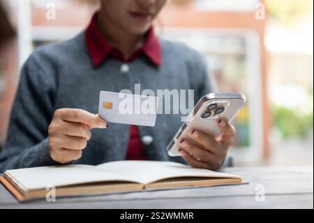 Ein zugeschnittenes Bild einer Frau, die ein Smartphone und eine Kreditkarte hält, während sie an einem Tisch im Freien sitzt, eine mobile Banking-App verwendet, Rechnungen online bezahlt, Stockfoto
