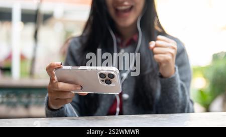 Ein zugeschnittenes Bild einer glücklichen asiatischen Frau trägt Ohrhörer, schaut sich einen Film an und spielt ein mobiles Spiel auf ihrem Smartphone, während sie draußen sitzt. peo Stockfoto