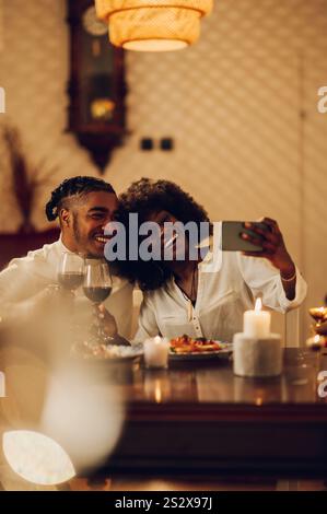 Verliebtes afroamerikanisches Paar feiert zu Hause mit einem romantischen Abendessen und Rotwein, während es ein Selfie mit einem Smartphone macht. Es macht Spaß, tim auszugeben Stockfoto