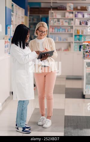 Die ganze Länge einer multikulturellen professionellen Apothekerin, die in der Drogerie steht und mit der Tablette in den Händen steht, mit ihrem älteren Patienten steht und diskutiert Stockfoto