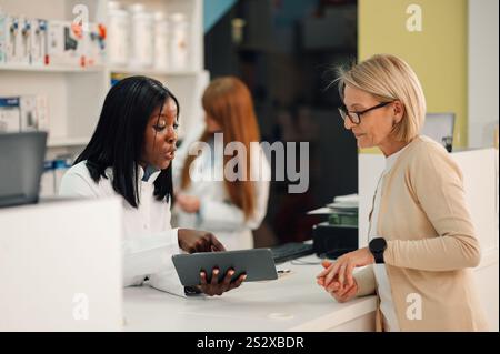 Professionelle multikulturelle Apothekerin, die in der Apotheke mit Tablette in den Händen steht und eine Seniorin konsultiert. Diverse Pharm Stockfoto