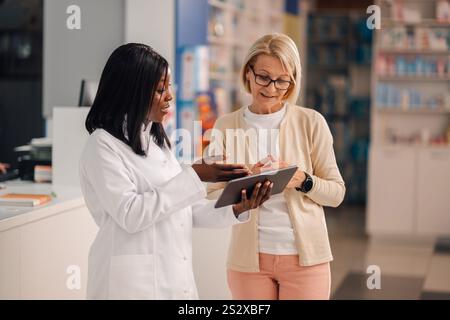 Professionelle weibliche multikulturelle Apotheke Ladenarbeiterin mit Tablette in den Händen, die gesundheitliche Bedenken mit älteren Frau diskutieren. Verantwortliche interrassische dru Stockfoto