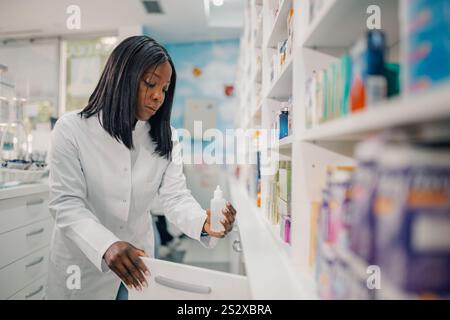 Porträt einer professionellen multikulturellen Apothekerin, die neben dem Schließfach steht, mit einer Flasche Medizin in den Händen und sie ansieht. Diverse Drogerien wo Stockfoto