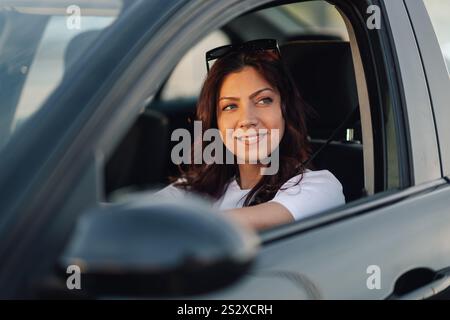 Porträt einer trendigen, lächelnden jungen Frau, die auf dem Vordersitz sitzt und beim Autofahren die Hände am Lenkrad hält. Nahaufnahme Stockfoto