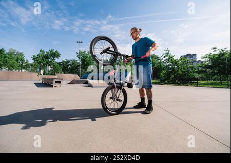 Ein urbaner, reifer Subkulturtyp steht mit seinem bmx in einem Skatepark und überprüft ihn. Ein Mann mittleren Alters mit einem grauen Pferdeschwanz posiert in einem Ska Stockfoto