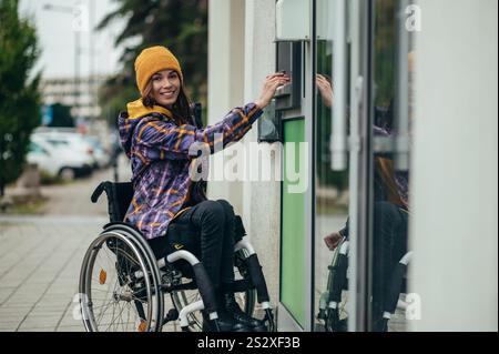 Junge schöne Frau mit Behinderung, die einen Rollstuhl benutzt, indem sie Kreditkarte und geldautomat benutzt, während sie Geld abhebt Stockfoto