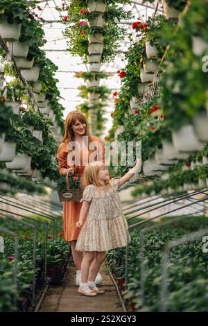 Die ganze Länge der jungen Mutter trägt Korb mit blühenden Blumen, während ihre entzückende kleine Tochter im Blumenkleid Blumen zum Kauf bei gr Stockfoto