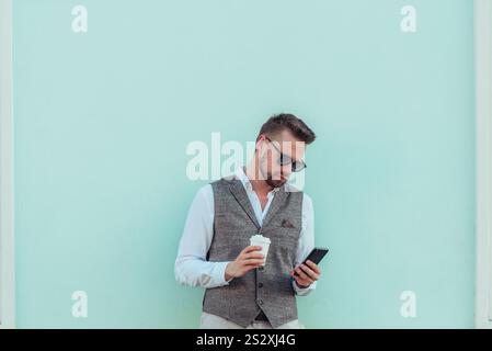 Aufnahme eines gut gekleideten Geschäftsmannes, der ein Smartphone benutzt und vor einer mintfarbenen Wand Kaffee trinkt Stockfoto