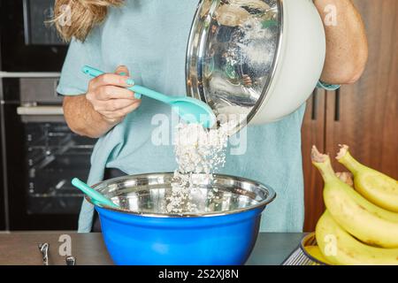 Eine Person gießt Mehl aus einer Metallschüssel in eine blaue Mischschüssel und bereitet die Zutaten für das Backen in der Küche vor. Stockfoto