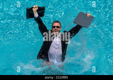 Ein lustiger, überraschter Geschäftsmann im Anzug mit einem Laptop im Swimmingpool. Verrückter, aufgeregter Geschäftsmann im Sommerurlaub. Aufgeregter Geschäftsmann in Neoprenanzug aufgeregt Stockfoto
