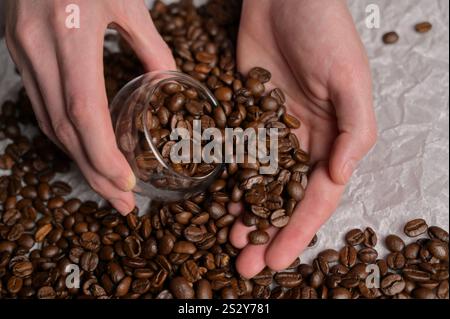 Frauenhände pflücken frische aromatisch geröstete Kaffeebohnen in ein kleines Glas, Handwerkspapier Hintergrund. Arabica- und Robusta-Mischung Stockfoto