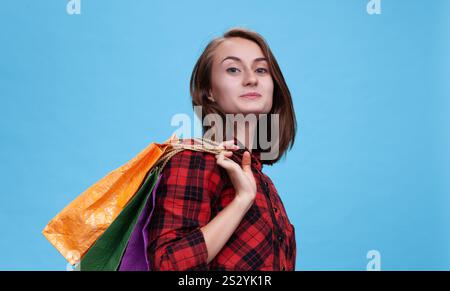 Eine fröhliche junge Frau steht selbstbewusst vor blauem Hintergrund und zeigt ihre pulsierenden Einkaufstaschen. Sie verkörpert die Freude der Handelssaison. Stockfoto
