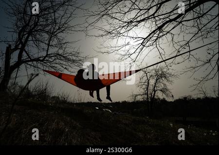 Silhouette von zwei Personen, die in einer orangen Hängematte zwischen blattlosen Bäumen liegen. Friedliche Outdoor-Szene. Stockfoto