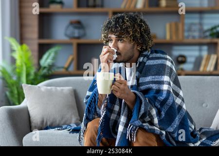 Kranker Mann mit Erkältung sitzt zu Hause auf der Couch, bedeckt mit einer Decke und mit einer Serviette in der Hand. Ein Mann mit einer Erkältung und einem heißen Getränk in der Hand hat eine laufende Nase. Stockfoto