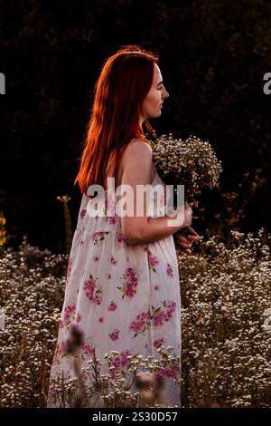 Ein rothaariges Mädchen steht in einem Gänseblümchenfeld, hält einen Blumenstrauß und blickt nachdenklich zur Seite Stockfoto