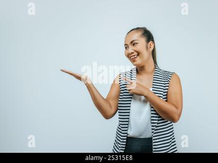 Porträt einer asiatischen Frau über isoliertem Hintergrund im Atelier mit leerem Raum. Lächelt beiseite, zeigt mit der Hand und zeigt mit dem Finger. Stockfoto