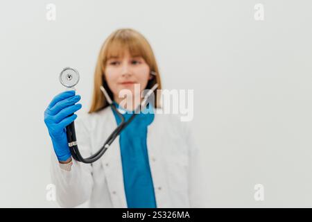 Eine Ärztin in einem weißen Mantel und blauen Handschuhen, die ein Stethoskop in Richtung Kamera hält. Medizinisches Fachpersonal im klinischen Umfeld Stockfoto